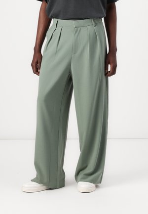 Pantalones de pierna ancha en un tejido verde apagado, con pliegues, una cintura alta y bolsillos laterales, combinados con zapatillas blancas sobre un fondo liso.