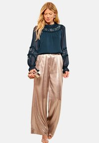 Tumši teal blūze ar ziedu izšuvumiem un volānu apkakli, saskaņota ar spīdīgām, platām, gaiši zelta biksēm. Modelis tur sudraba rakstāmmašīnu.