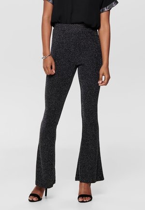 Femme portant un pantalon noir évasé pailleté taille haute avec des talons ouverts noirs, associé à un haut noir avec des manches bordées de métal.