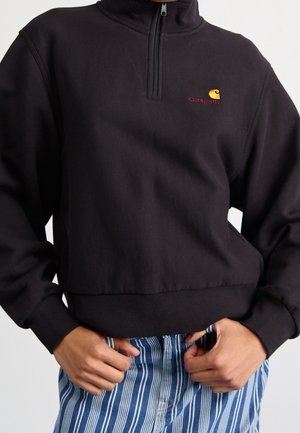 Osoba ubrana w czarną bluzę Carhartt z ćwiartkowym zamkiem i ściągaczami na mankietach oraz u dołu, w połączeniu z niebiesko-białymi pionowo paskowanymi spodniami.