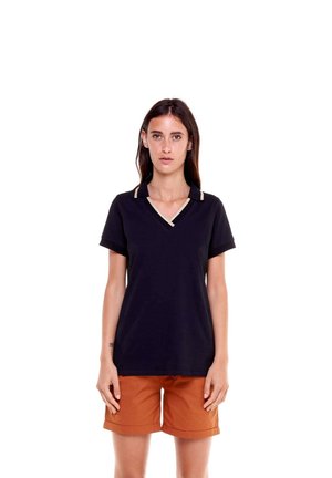 Une femme porte un haut noir à manches courtes et à encolure en V, bordé de beige, ainsi qu’un short orange rouille, sur fond blanc.
