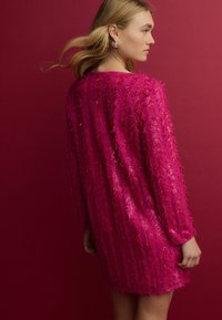 Fuchsia jurk met lange mouwen, bedekt met getextureerde franjes en pailletten. Gladde stof, knielange ontwerp, tegen een rode achtergrond.
