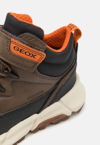 Baskets brunes et noires avec des touches d'orange, présentant un dessus texturé, un col rembourré et un marquage "GEOX" bien visible. Semelle en caoutchouc.