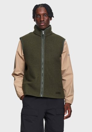Homem com cabelo curto e enrolado, vestindo um colete verde-oliva com fecho zip sobre uma camisola bege de mangas compridas e calças cargo pretas, de pé contra um fundo simples.