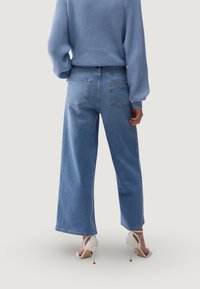 Laiade säärega sinised teksad, valmistatud denimist, kõrge vöökohaga ja tagataskutega, kombineeritud helesinise kootud kampsuni ja valgete kõrgete kontsadega.