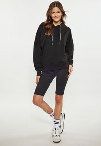 Felpa nera oversize con maniche lunghe, dettagli con cordoncino, abbinata a pantaloni ciclistici neri e sneakers bianche con strisce nere e rosse.