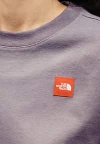 Κοντινή λήψη ατόμου που φοράει μωβ φούτερ με στρογγυλή λαιμόκοψη, με μικρό κόκκινο τετράγωνο έμβλημα που έχει το λευκό λογότυπο The North Face στο στήθος.