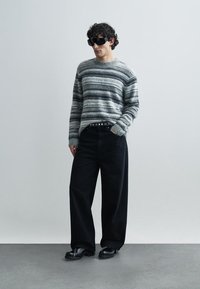 Grau gestreifter Strickpullover, schwarze Hosen mit weitem Bein und schwarze Slipper mit strukturierter Oberfläche und einer lockeren Silhouette. Dunkle Sonnenbrille.