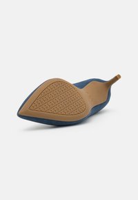 Chaussure à talon haut pointu bleu marine vue de la semelle avec une semelle texturée beige et un talon fin sur fond blanc.