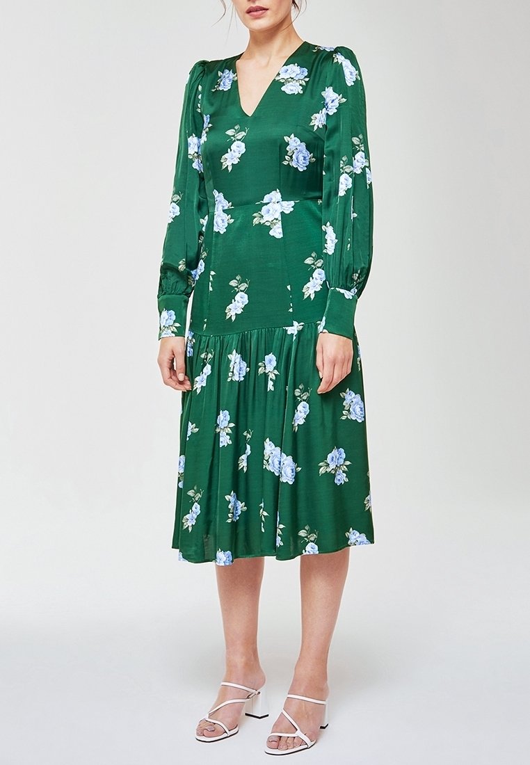 Femme portant une robe midi verte à manches longues et col en V, avec un imprimé floral bleu et blanc, et des sandales blanches à talons, debout devant un fond uni.