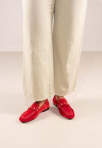 Mocasines de ante rojo con tachuelas decorativas y detalles de costura, combinados con pantalones de pierna ancha beige claro. Suela plana y diseño de punta redondeada.
