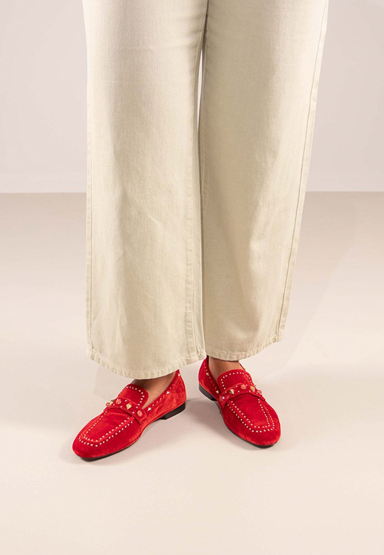 Mocasines de ante rojo con tachuelas decorativas y detalles de costura, combinados con pantalones de pierna ancha beige claro. Suela plana y diseño de punta redondeada.