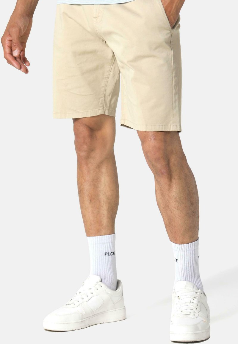 Shorts en coton beige avec poches, coupe droite et ourlet au-dessus du genou. Associés à des baskets blanches et des chaussettes blanches côtelées.