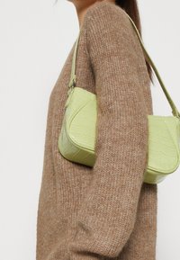 Zielona torebka z tłoczeniem imitującym skórę krokodyla, o zaokrąglonym kształcie i długim pasku, w zestawieniu z brązowym, teksturowanym swetrem.