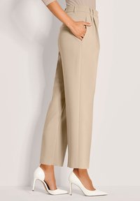 Vrouw in beige op maat gemaakte broek en witte hoge hakken, staand met één hand in de zak tegen een effen achtergrond.