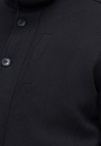 Czarna kurtka z mieszanki wełny z wysokim kołnierzem, z dwoma dużymi czarnymi guzikami i subtelną fakturą powierzchni. Dyskretne, ale zauważalne detale kieszeni.