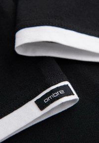 Černá látka s bílým lemem ukazuje detail manžety rukávu, na které je malá černá nálepka s bílým textem "ombre".