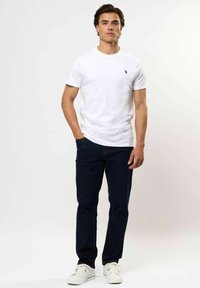 Férfi áll szembe, fehér rövidujjú pólót, sötétkék farmert és fehér sportcipőt viselve, kezét részben a zsebében tartva.