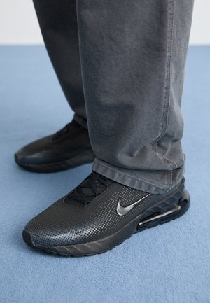 Baskets Nike noires portées avec un pantalon gris foncé sur un sol recouvert d'une moquette bleu clair.