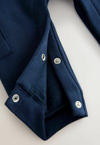 Tmavomodré bavlnené nohavice s patentkami na spodnej časti, s mäkkou vnútornou podšívkou a prešitými švami. Kovové patentky viditeľné na leme.