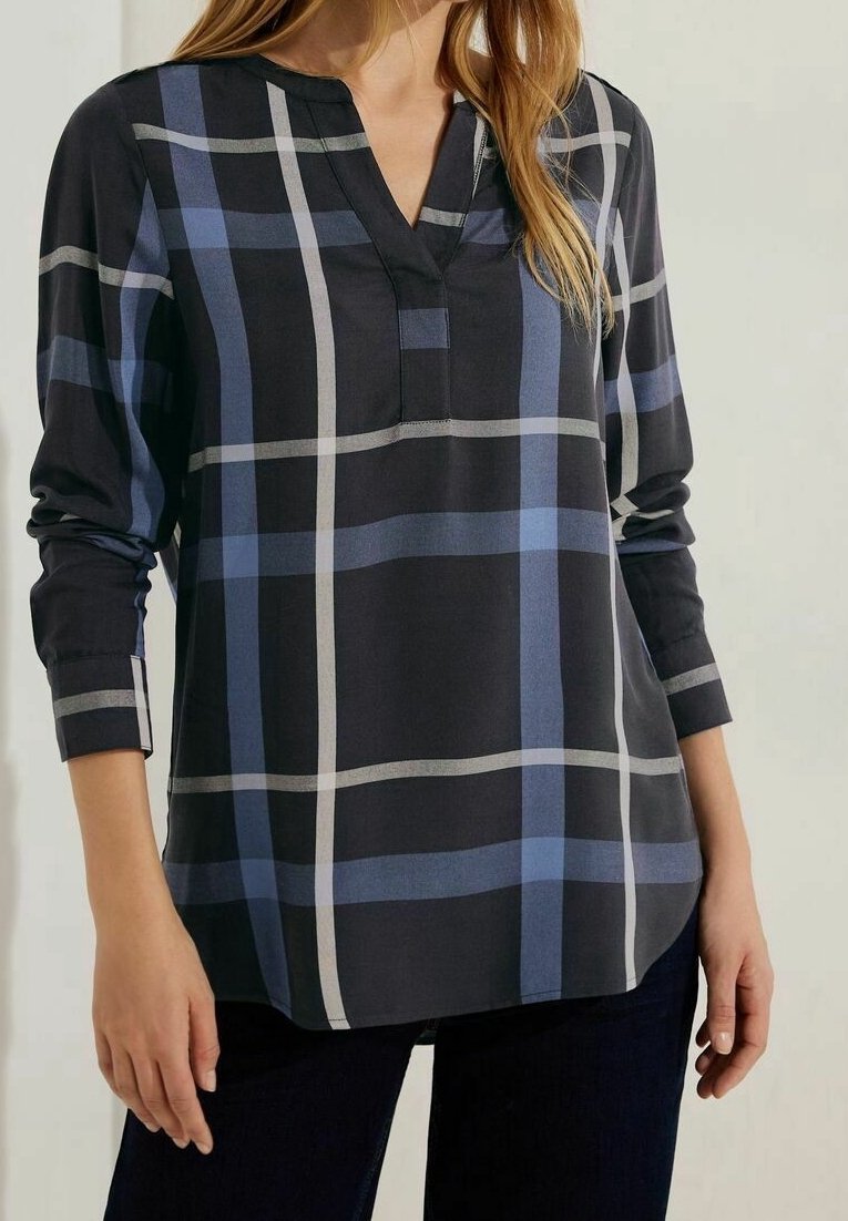 Femme portant un chemisier noir à manches longues avec un grand motif à carreaux bleu et gris et un col en V, assorti à un pantalon foncé.