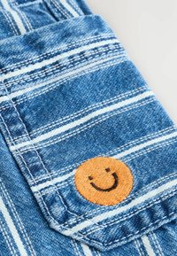 Denim z niebiesko-białym paskowanym wzorem, z kieszonką z pomarańczowym haftowanym uśmiechniętą buźką. Tekstura jest gładka, casualowy design.