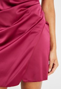 Robe en satin fuchsia avec un tissu drapé, présentant un détail de couture latérale et un ourlet légèrement inégal. Texture lisse avec un léger éclat.