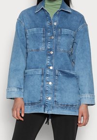 Denim jacket in light blue with two chest pockets, silver buttons, and a relaxed fit. Features a collared neckline and cuffed sleeves.