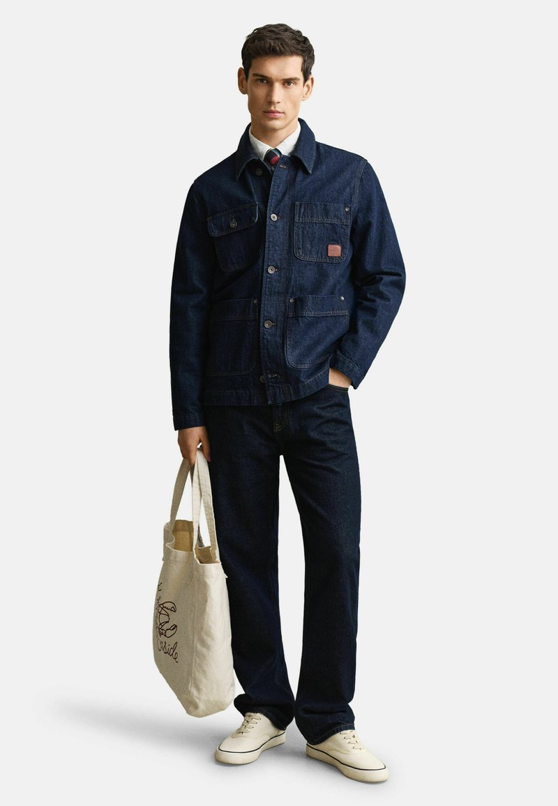 Uomo con giacca e pantaloni di denim scuro, sneaker bianche, che tiene una borsa tote beige con design a granchio, in piedi davanti a uno sfondo bianco.