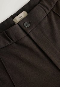 Pantaloni su misura di colore marrone scuro con chiusura a un bottone, parte anteriore plissettata e etichetta con la scritta "NEXT Slim Fit". Il tessuto ha una texture liscia.