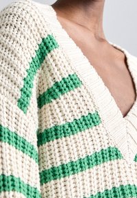 Pull tricoté couleur crème avec des rayures horizontales vertes, doté d'un col en V profond et d'un tissu texturé. Détails en côtes proéminents.