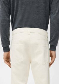 Pantalones de mezclilla blancos con un diseño ajustado, que cuentan con bolsillos traseros y una cinturilla lisa. Se ve parcialmente una camiseta de manga larga de color gris oscuro.