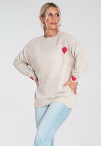 Beżowy sweter z dzianiny o luźnym kroju, z czerwonymi akcentami w kształcie serca na klatce piersiowej i rękawach. Ściągacze na mankietach i u dołu. Pod spodem noszone dżinsy.