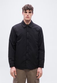 Νέος άνδρας με σγουρά μαλλιά, φοράει μαύρο σακάκι με κουμπιά και καφέ παντελόνι cargo, στέκεται μπροστά σε απλό ανοιχτό φωτεινό φόντο.