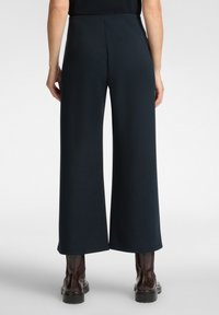 Kalhoty s širokými nohavicemi ve tmavě modré látce, s hladkou texturou a designem s vysokým pasem. Doplňují je hnědé boty s robustní podrážkou.