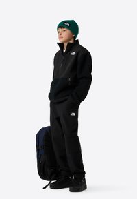 Crna dukserica s patentnim zatvaračem i teksturiranim donjim dijelom, u kombinaciji s crnim hlačama. Zelena pletena kapa i crne tenisice upotpunjuju odjevnu kombinaciju.