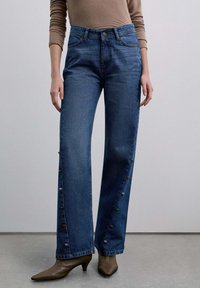 Vaqueros de pierna ancha en azul hechos de mezclilla con cierre de botones, bolsillos laterales y adornos decorativos de colores a lo largo de las costuras exteriores.