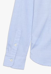 Camicia azzurro chiaro, con bottoni e una texture liscia, dotata di maniche lunghe e polsini arrotondati con bottoni bianchi e dettagli cuciti.