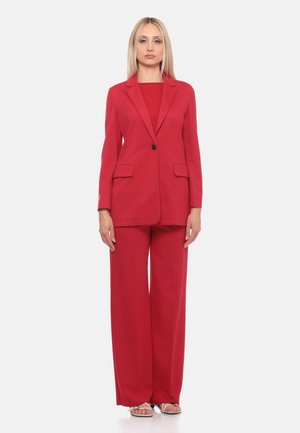 Donna in piedi rivolta in avanti che indossa un blazer rosso, pantaloni larghi coordinati e sandali su uno sfondo chiaro e uniforme.