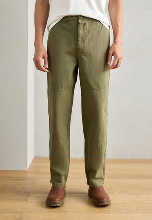 Homme portant un pantalon droit vert olive, des bottes en cuir marron et une chemise blanche, debout sur un sol en bois à l'intérieur.