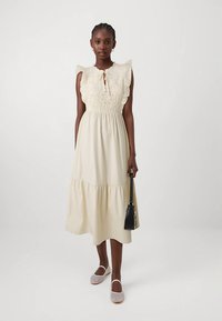 Krēmkrāsas kleita ar volānu izgriezumu, mežģīņu detaļām un kārtotu svārku. Kombinēta ar nelielu melnu rokassomu un austiem apaviem.