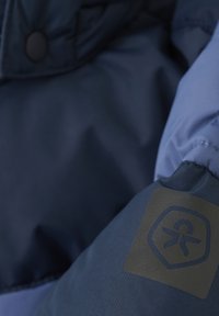 Mörkblå isolerad jacka med en slät yta, med en svart logopatch på ärmen och tryckknappdetaljer.