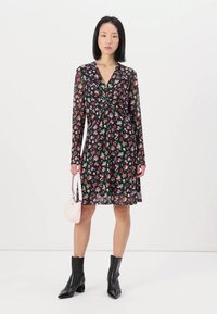 Robe noire à fleurs avec des manches longues, présentant un détail noué à l'avant, complétée par un sac à main pastel et des bottines noires brillantes.