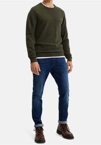 Svetle olivově zelený svetr textury s kulatým výstřihem, nošený přes bílé tričko. Ve spojení s modrými džínami a hnědými šněrovacími botami.