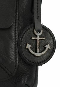 Detail van een zwart leren tas met een gestructureerd oppervlak en een ronde label met een zilveren ankerembleem en geperst tekst.