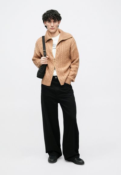 Gaiši brūns adīts džemperis ar augstu apkakli un rāvējslēdzēju, valkāts pār baltu kreklu. Melnas plašas bikšu kājas un melni apavi.