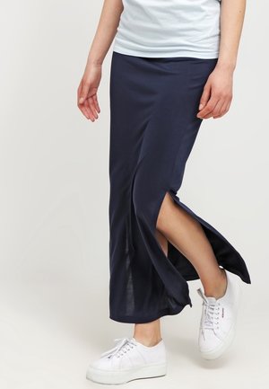 Persoon die een marineblauwe midi-rok met een zijsplit draagt en witte platform sneakers, staand met één voet iets opgetild.