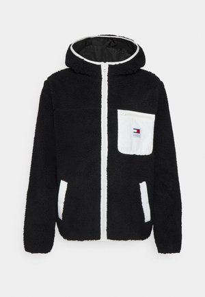 Musta fleecehuppari, jossa valkoinen vetoketju, valkoiset reunukset taskuissa ja valkoinen vetoketjullinen rintatasku, jossa pieni logopatch.