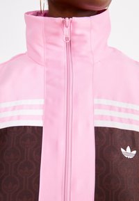 Rosa zip-up jacka med hög krage, med bruna textuerade paneler och vita tre-stripes detaljer, samt en logotyp på den nedre delen.