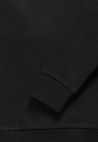 Tessuto felpa nera che mostra un primo piano del polsino e dell'orlo, con una texture a coste e una superficie liscia. Le cuciture minime sono visibili.